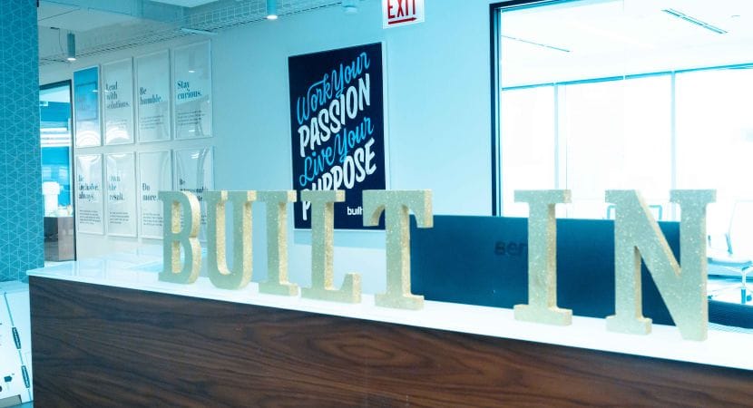 Photo of front desk with "Built In" block letters on countertop, and sign in background that reads "Work your passion, live your purpose"