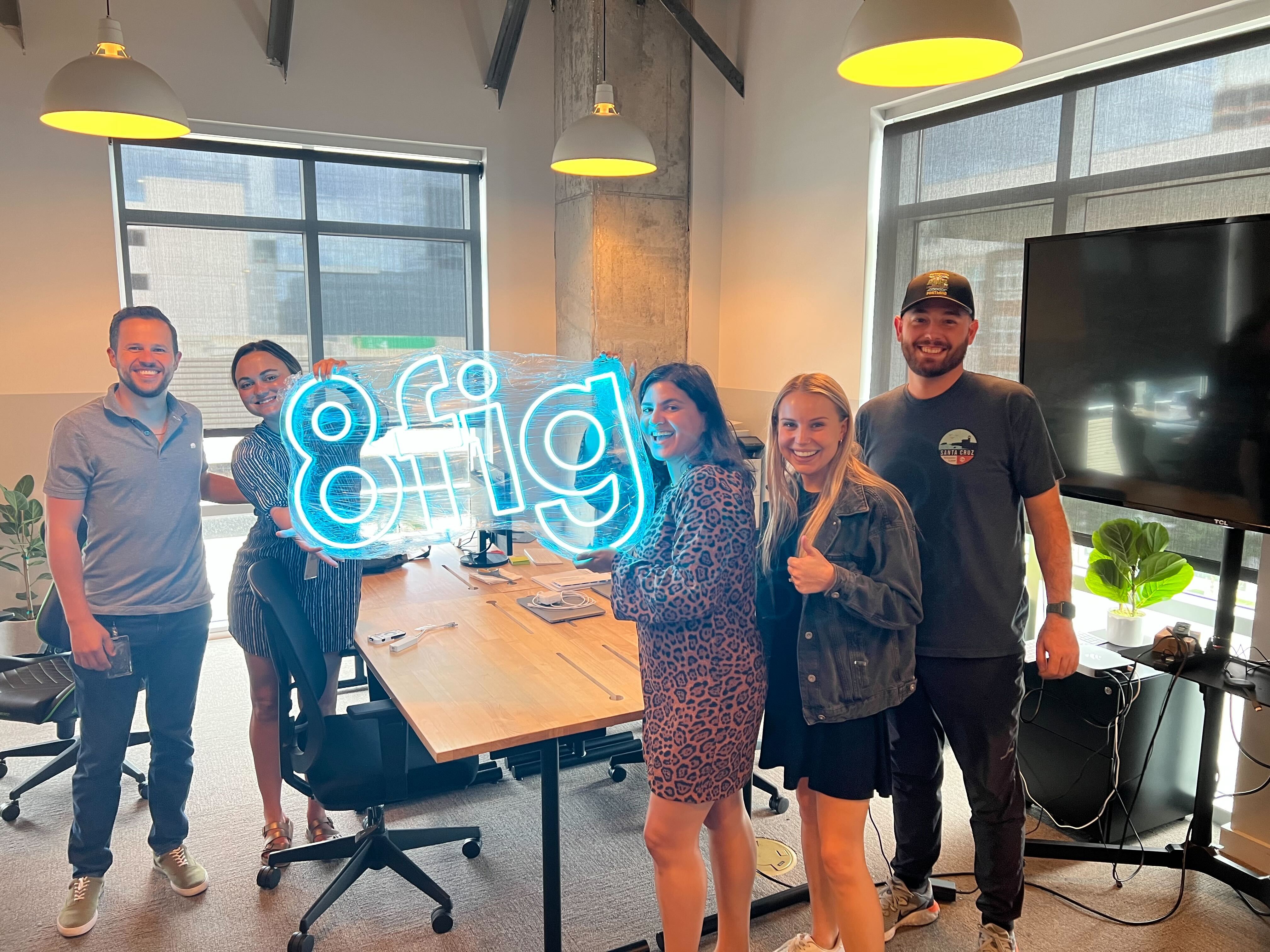 Small team inside of an office meeting room holding a large, lighted 8fig logo.