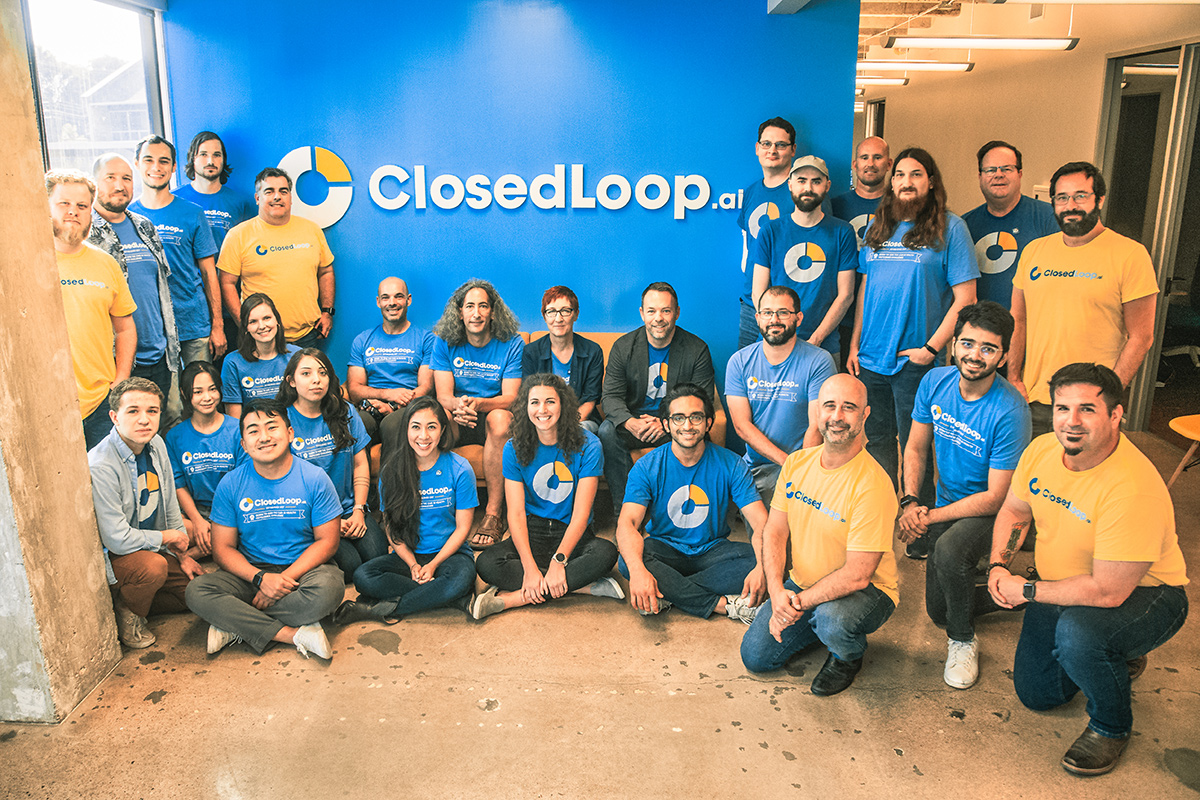 ClosedLoop team members wearing t-shirts with the company logo and the logo on a blue wall behind them