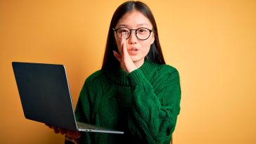 Young woman wearing glasses and holding a laptop with her hand by her mouth like she's telling a secret