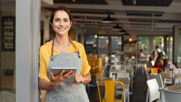 A woman in an apron leaning against the doorway of a restaurant, holding a tablet and smiling.