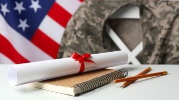 An American flag waves in the background. In the foreground, a military uniform jacket is hung on a chair. In front of it on a table is a rolled up diploma with a red ribbon tied around it sitting on top of a notebook, with some pencils placed next to it.