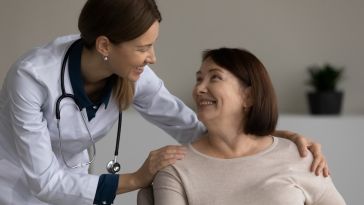 A woman doctor leans over a woman patient's shoulder to talk to her. The doctor touches the patient's shoulder and they are smiling at one another.