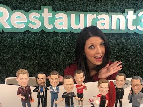 SVP of People Jill Burke sits at the front desk in Restaurant365’s office, with custom bobbleheads of company leaders in front of her and the company logo on the wall behind her.